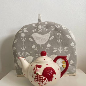 May include: White ceramic teapot with a red rooster design and white polka dots. The teapot has a red handle and spout. The teapot is sitting on a white surface with a gray and white tea cozy with a bird design.