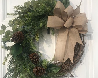 May include: A brown burlap bow with a green wreath made of pine, eucalyptus, and magnolia leaves. The wreath is decorated with pine cones. The wreath is hanging on a white door. The text "Autumn's Echo Shoppe" is written in green cursive at the bottom of the image.