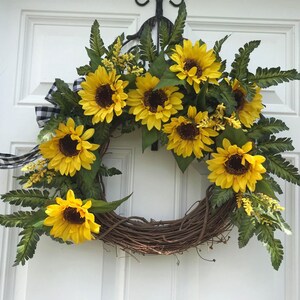 Pu&ograve; includere: Una ghirlanda di girasoli con nastro a quadri bianco e nero. La ghirlanda &egrave; fatta di vite e presenta diversi girasoli con foglie verdi.