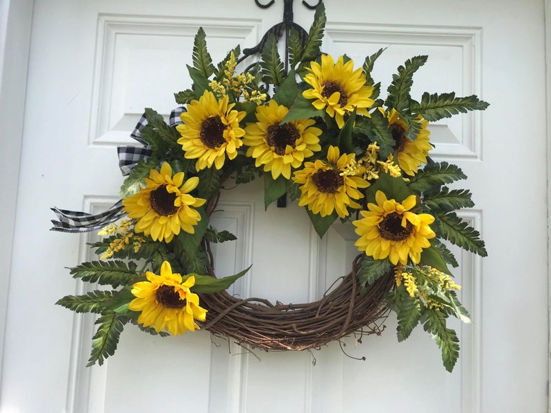 Pu&ograve; includere: Una ghirlanda di girasoli con nastro a quadri bianco e nero. La ghirlanda &egrave; fatta di vite e presenta diversi girasoli con foglie verdi.