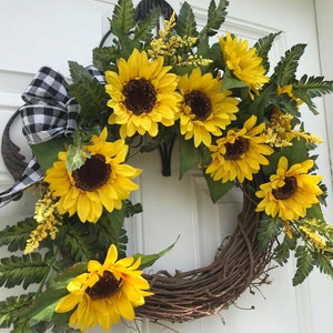 Pu&ograve; includere: Una ghirlanda di girasoli con un nastro a scacchi bianco e nero. La ghirlanda &egrave; fatta di vite e ha una variet&agrave; di foglie verdi e girasoli gialli.
