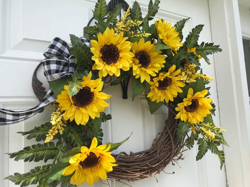 Pu&ograve; includere: Una ghirlanda di girasoli con un nastro a scacchi bianco e nero. La ghirlanda &egrave; fatta di vite e ha una variet&agrave; di foglie verdi e girasoli gialli.