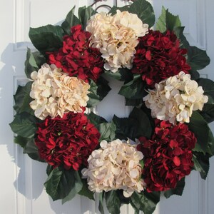Puede incluir: Una corona hecha de hortensias artificiales en tonos crema y burdeos. La corona est&aacute; decorada con hojas verdes y es adecuada para una puerta de entrada.