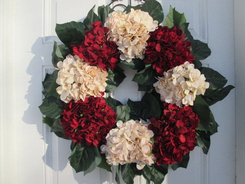 Puede incluir: Una corona hecha de hortensias artificiales en tonos crema y burdeos. La corona est&aacute; decorada con hojas verdes y es adecuada para una puerta de entrada.