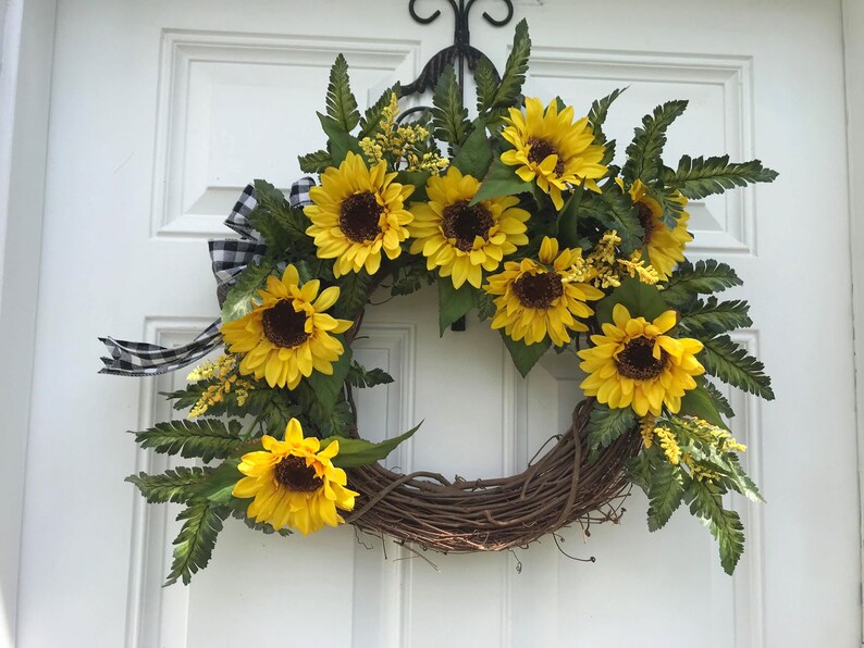 Pu&ograve; includere: Una ghirlanda di girasoli con un nastro a quadri bianco e nero. La ghirlanda &egrave; realizzata in vite e presenta una variet&agrave; di girasoli di diverse dimensioni e stadi di fioritura.