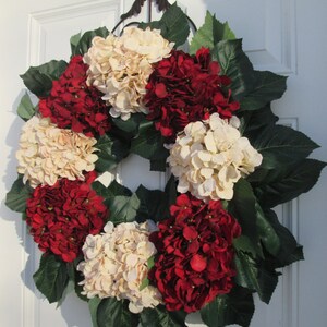 Puede incluir: Una corona hecha de hortensias artificiales en tonos crema y rojo burdeos. La corona est&aacute; decorada con hojas verdes y est&aacute; colgada en una puerta blanca.
