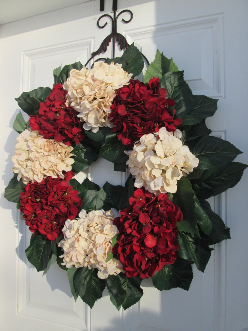 Puede incluir: Una corona hecha de hortensias artificiales en tonos crema y rojo burdeos. La corona est&aacute; decorada con hojas verdes y est&aacute; colgada en una puerta blanca.