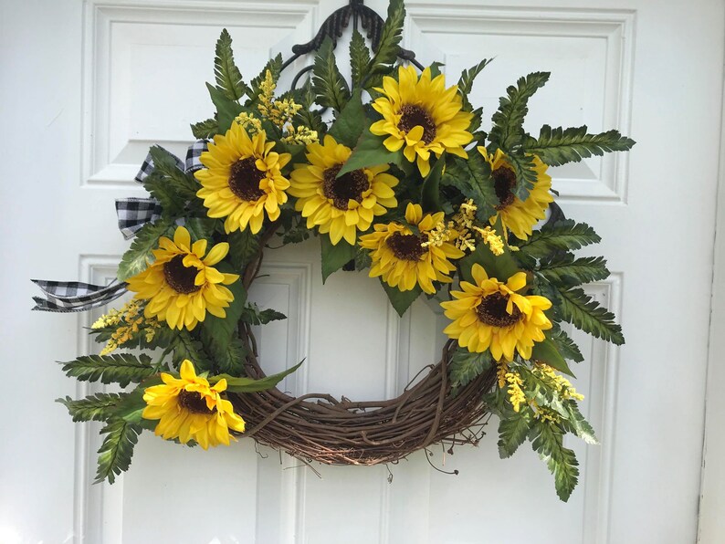 Pu&ograve; includere: Una ghirlanda di girasoli con un nastro a scacchi bianco e nero. La ghirlanda &egrave; realizzata in vite e presenta diversi girasoli con petali gialli e centri marroni. La ghirlanda &egrave; decorata con foglie di felce verdi.