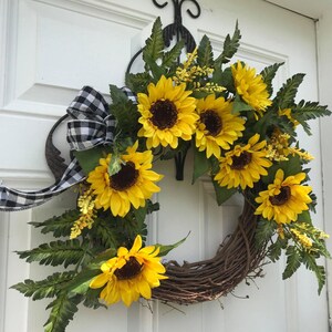 Pu&ograve; includere: Una ghirlanda di girasoli con un nastro a scacchi bianco e nero. La ghirlanda &egrave; fatta di vite e ha una variet&agrave; di girasoli di diverse dimensioni. La ghirlanda &egrave; appesa a una porta bianca.