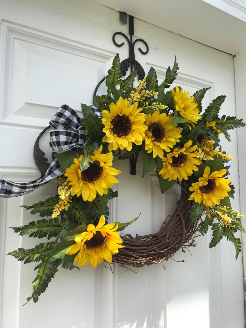 Pu&ograve; includere: Una ghirlanda di girasoli con un nastro a scacchi bianco e nero. La ghirlanda &egrave; fatta di vite e ha una variet&agrave; di girasoli di diverse dimensioni. La ghirlanda &egrave; appesa a una porta bianca.