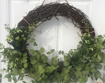 May include: A green leafy wreath made of eucalyptus and other greenery, with a brown vine base. The wreath is hanging on a white door.
