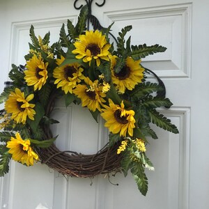 Pu&ograve; includere: Una ghirlanda di girasoli con una base di vite marrone, appesa a una porta bianca. La ghirlanda &egrave; decorata con girasoli gialli e felci verdi.