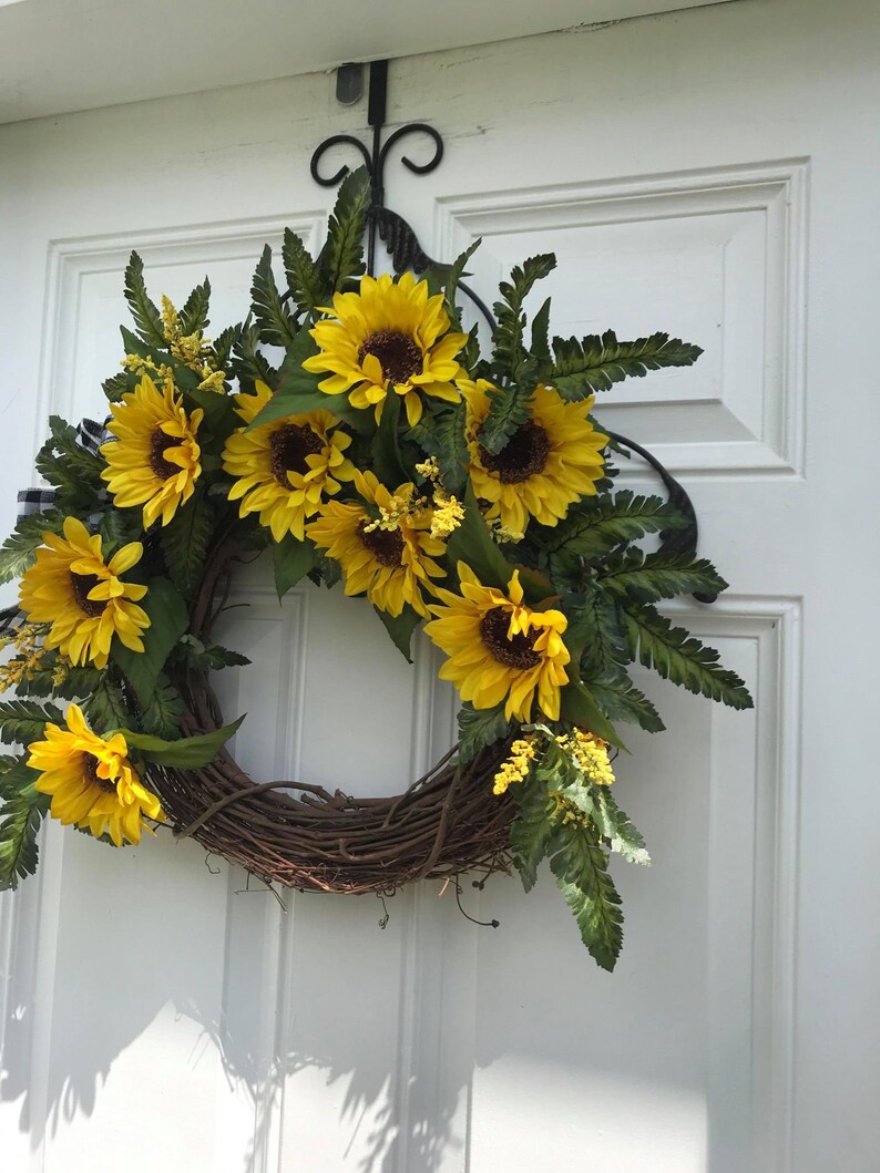 Pu&ograve; includere: Una ghirlanda di girasoli con una base di vite marrone, appesa a una porta bianca. La ghirlanda &egrave; decorata con girasoli gialli e felci verdi.