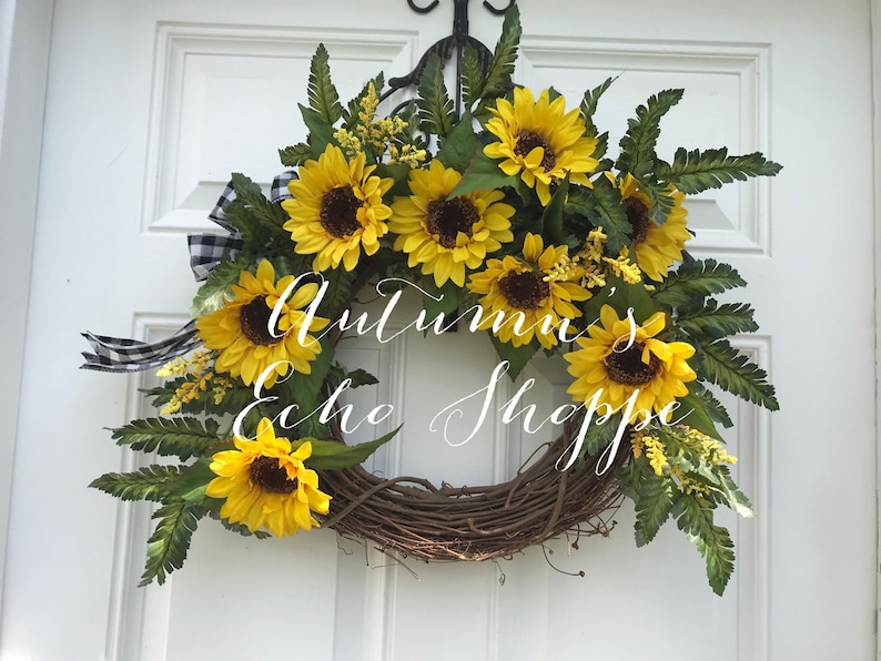 Pu&ograve; includere: Una ghirlanda di girasoli con un nastro a scacchi bianco e nero. La ghirlanda &egrave; fatta di vite e ha una variet&agrave; di foglie verdi. Il testo "Autumn's Echo Shoppe" &egrave; scritto in corsivo bianco sulla ghirlanda.