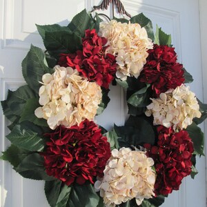 Puede incluir: Una corona hecha de hortensias artificiales en tonos crema y rojo burdeos. La corona est&aacute; decorada con hojas verdes y est&aacute; colgada en una puerta blanca.