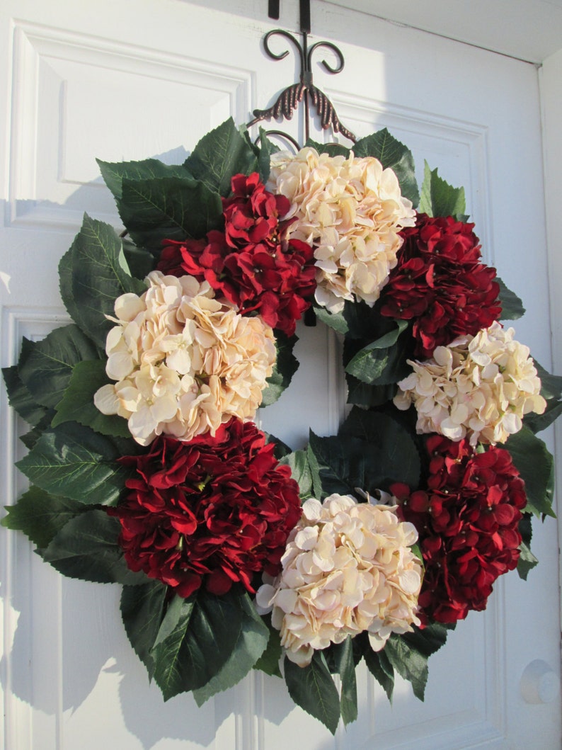 Puede incluir: Una corona hecha de hortensias artificiales en tonos crema y rojo burdeos. La corona est&aacute; decorada con hojas verdes y est&aacute; colgada en una puerta blanca.