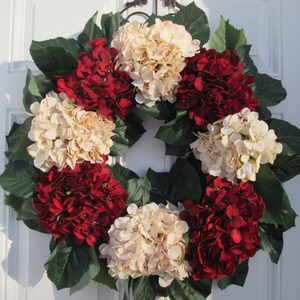 Puede incluir: Una corona hecha de hortensias artificiales en tonos crema y burdeos. La corona est&aacute; decorada con hojas verdes y es adecuada para una puerta de entrada.
