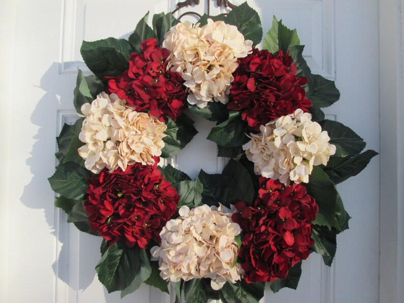 Puede incluir: Una corona hecha de hortensias artificiales en tonos crema y burdeos. La corona est&aacute; decorada con hojas verdes y es adecuada para una puerta de entrada.