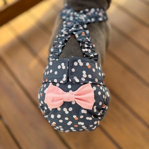 Puede incluir: Un perro con un pañal azul marino con lunares blancos y naranjas y un lazo rosa claro. El pañal tiene correas ajustables y un arnés de lunares a juego.