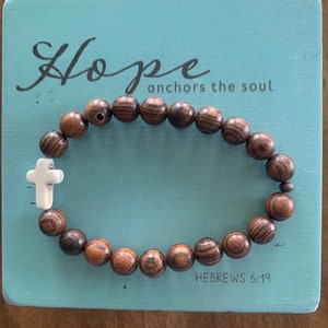 May include: A wooden beaded bracelet with a white cross charm. The bracelet is on a blue background with the text "Hope anchors the soul" and "HEBREWS 6:19".