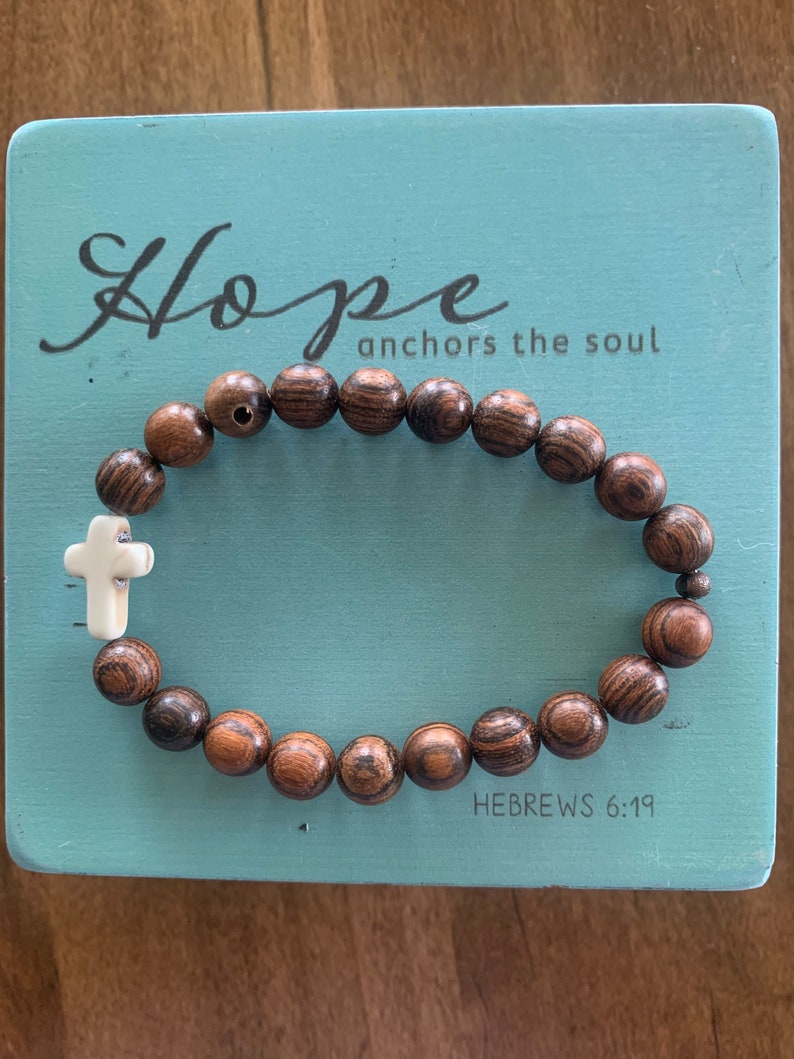 May include: A wooden beaded bracelet with a white cross charm. The bracelet is on a blue background with the text "Hope anchors the soul" and "HEBREWS 6:19".