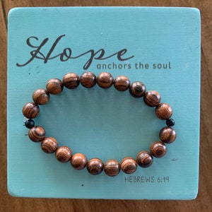 May include: A wooden beaded bracelet with black beads interspersed throughout. The bracelet is on a light blue background with the text "Hope anchors the soul" and "HEBREWS 6:19".