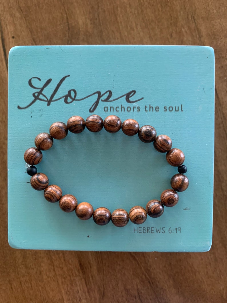 May include: A wooden beaded bracelet with black beads interspersed throughout. The bracelet is on a light blue background with the text "Hope anchors the soul" and "HEBREWS 6:19".