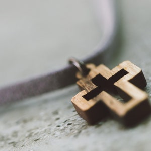 May include: A wooden cross pendant on a light purple cord. The cross is a simple design with a slightly distressed finish.