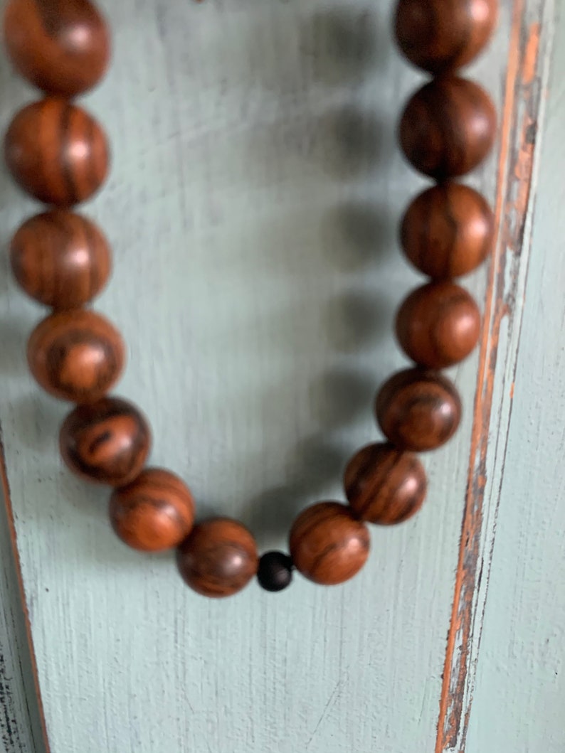 May include: A brown wooden beaded necklace with a black bead accent. The beads are round and have a natural wood grain pattern.