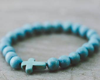 May include: A blue beaded bracelet with a turquoise cross charm. The bracelet is made of small, round beads and is strung on a thin, elastic cord.