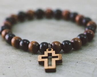 May include: A wooden cross charm hangs from a brown and black beaded bracelet.