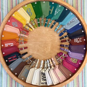 May include: A circular collection of numerous rectangular luggage tags in a wide spectrum of colors, including red, orange, yellow, green, blue, purple, pink, and metallic tones. Each tag features a silver-toned buckle and numbered labels.