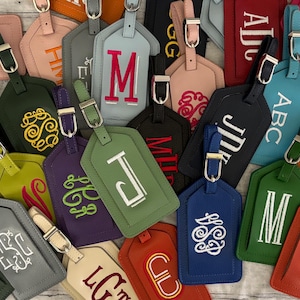 May include: A collection of rectangular luggage tags in various colors including pink, green, blue, orange, black, and grey, each featuring a silver metal buckle and personalized embroidered monograms or initials in contrasting thread colors.