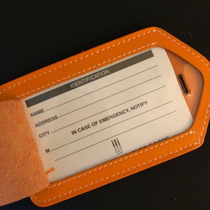 May include: An orange leather luggage tag with white stitching and a clear plastic window. The white identification card inside features the text IDENTIFICATION, NAME, ADDRESS, CITY, M, and IN CASE OF EMERGENCY, NOTIFY, along with a small logo.