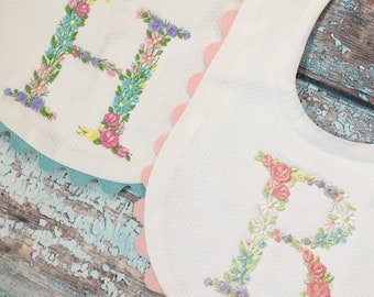 May include: Two white baby bibs with floral embroidered letters, one with an "H" and the other with a "B". The bibs have scalloped edges in light pink and light blue. The letters are made of colorful flowers.