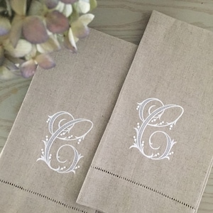 May include: Two beige linen hand towels with a white embroidered letter "C" and decorative floral accents. Each towel has a stitched border along the bottom edge. The towels are folded and arranged side by side.