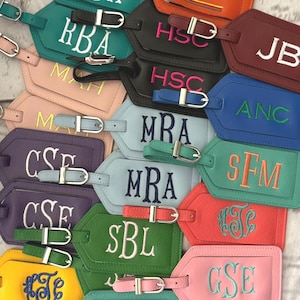 May include: A collection of rectangular luggage tags in various colors including orange, teal, black, burgundy, blue, purple, green, yellow, and pink. Each tag features a silver metal buckle strap and embroidered monogram letters or initials.