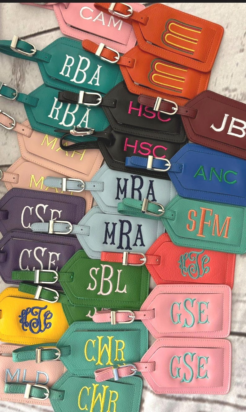 May include: A collection of rectangular luggage tags in various colors including orange, teal, black, burgundy, blue, purple, green, yellow, and pink. Each tag features a silver metal buckle strap and embroidered monogram letters or initials.