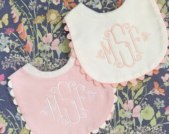 May include: Two pink and white scalloped bibs with embroidered white monograms. The bibs are on a floral patterned background.