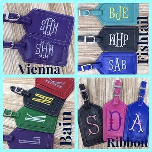 May include: A collection of rectangular luggage tags in various colors including purple, teal, black, blue, red, green, and pink. Each tag features embroidered monograms and includes a silver-toned metal buckle strap for attachment to bags.