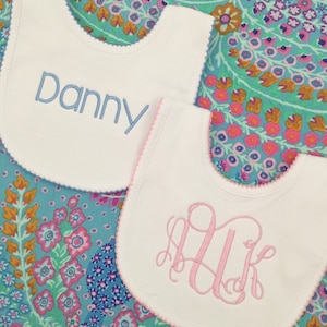 May include: Two white baby bibs with embroidered designs. One bib has the name "Danny" embroidered in blue thread. The other bib has a pink monogram embroidered in the center.