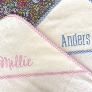 May include: Two white hooded towels with embroidered names. One towel features pink gingham trim and the name Millie in pink script. The other towel has blue gingham trim and the name Anders in blue block letters. Both are made of soft terry cloth.