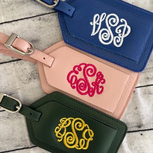 May include: Three rectangular luggage tags with silver-toned metal buckles. The top tag is blue with white embroidered monogram letters. The middle tag is light pink with hot pink embroidery. The bottom tag is dark green with yellow embroidery.