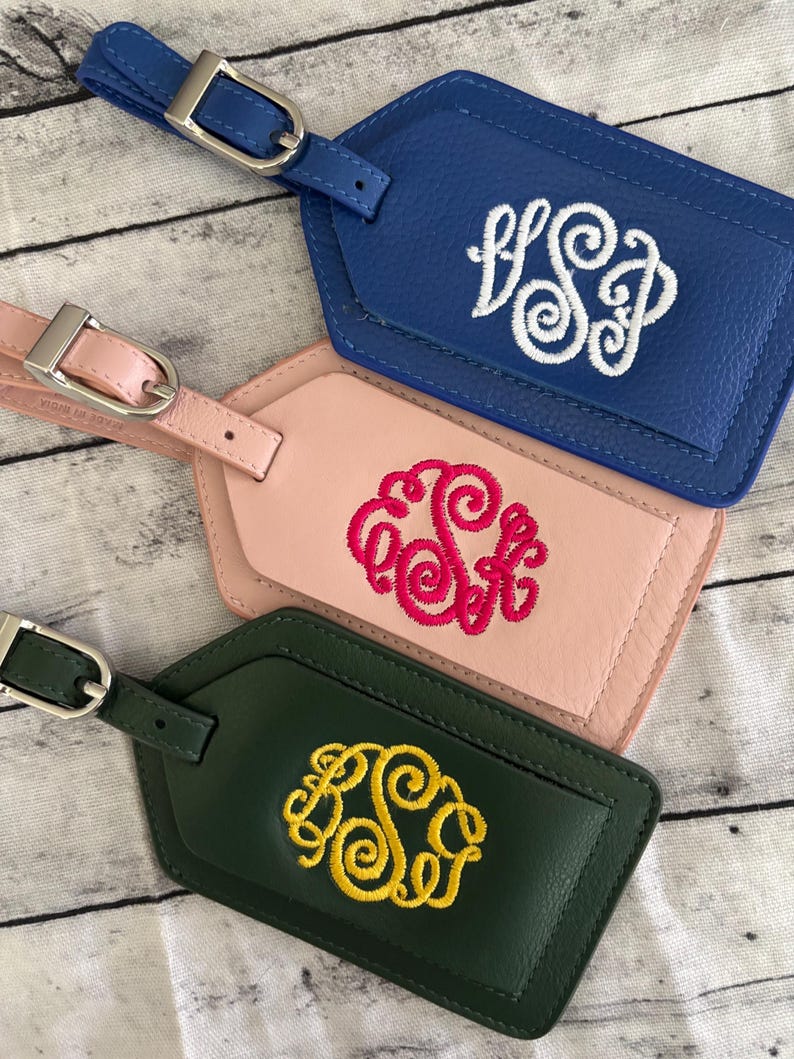 May include: Three rectangular luggage tags with silver-toned metal buckles. The top tag is blue with white embroidered monogram letters. The middle tag is light pink with hot pink embroidery. The bottom tag is dark green with yellow embroidery.