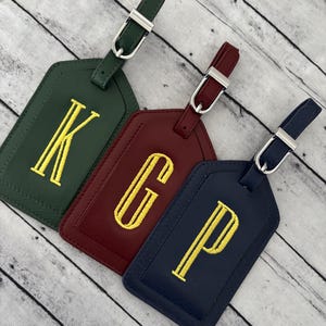 May include: Three rectangular luggage tags in dark green, burgundy, and navy blue. Each tag features a single embroidered yellow letter, K, G, or P, and includes a matching strap with a silver-toned metal buckle for secure attachment to travel bags.