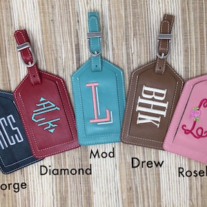 May include: Five rectangular luggage tags with adjustable straps and silver buckles. The tags are dark blue with white ACS, red with teal Diamond, teal with pink Mod L, brown with white BHK, and pink with a floral Rosebud L monogram embroidery.