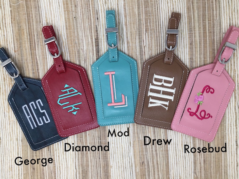 May include: Five rectangular luggage tags with adjustable straps and silver buckles. The tags are dark blue with white ACS, red with teal Diamond, teal with pink Mod L, brown with white BHK, and pink with a floral Rosebud L monogram embroidery.