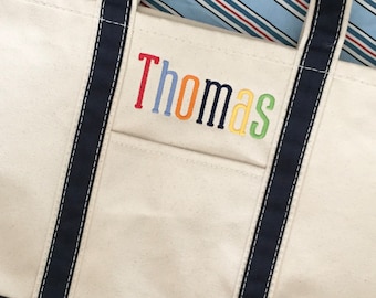 May include: A white canvas tote bag with navy blue straps and a rainbow embroidered name "Thomas".