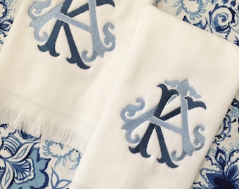 May include: Two white rectangular hand towels featuring a fringed bottom edge and an embroidered monogram in shades of dark and light blue. The monogram consists of the letters A and K intertwined in a decorative, ornate script style.