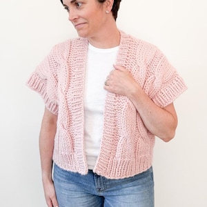 Puede incluir: Un cárdigan de punto rosa con mangas cortas y un patrón de punto trenzado. El cárdigan está abierto por delante y tiene un corte holgado.